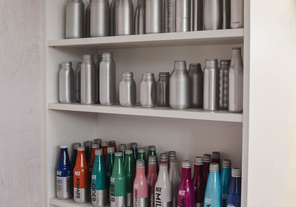 A collection of insulated stainless steel bottles and can-shaped drink containers arranged on white shelves. The top two shelves display mostly silver and gray bottles, while the bottom shelf features a row of insulated bottles in multiple colors including blue, orange, teal, green, pink, and purple.
