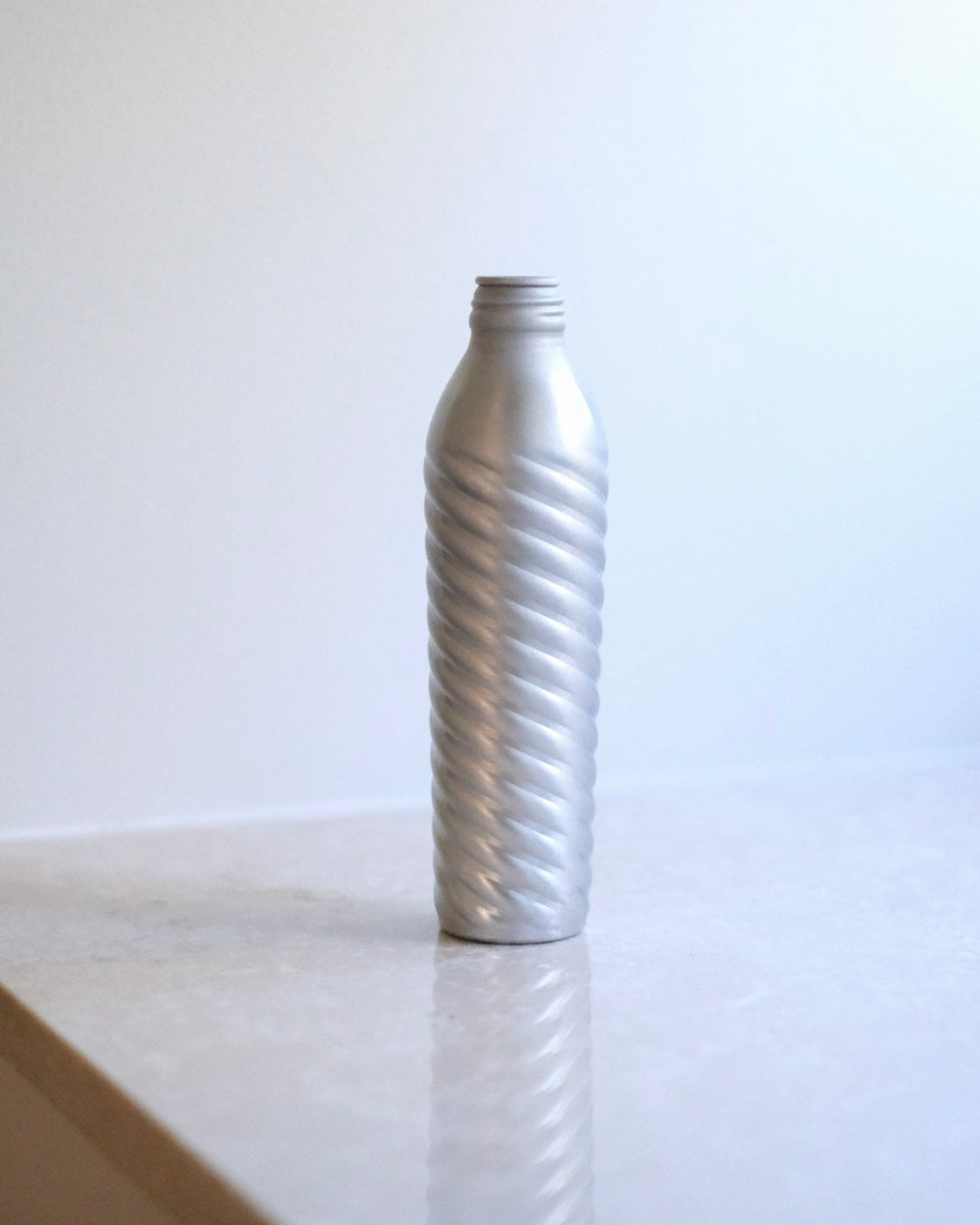 A silver aluminum bottle with a diagonal spiral ribbed pattern, standing on a light marble countertop against a white wall.