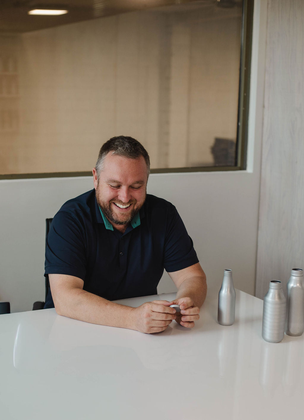 A smiling man in a navy polo shirt sits at a white table with several silver aluminum bottles nearby in an indoor office or studio setting.